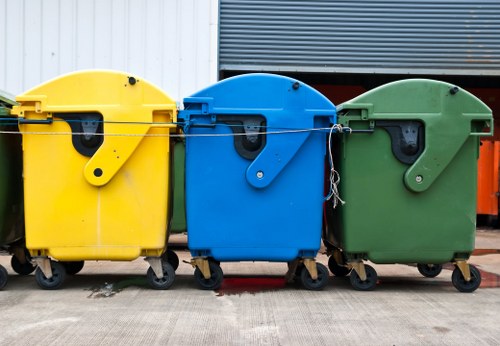 Three large wheeled rubbish bins are positioned side by side on a concrete surface, with the yellow bin on the left, a blue bin in the middle, and a green bin on the right. Each bin has a rounded lid with a small black handle, and they are constructed from durable plastic with a textured surface. The yellow bin features a smooth, matte finish; the blue bin has a slightly glossy surface; and the green bin displays a matte finish. The bins are set against a backdrop of a metal structure, with a corrugated metal wall visible above, suggesting an industrial or commercial environment typical for waste management in Maida Vale. The wheels and hinges of each bin are visible at the base, and the bins are positioned on a paved, slightly worn surface, with some minor debris and stains. The overall scene captures a standard setup for rubbish collection or waste removal services provided by businesses like Business Waste Removal Maida Vale, highlighting their focus on container-based waste management solutions in the area.