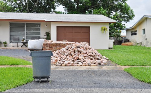 In the driveway of a residential property, a large pile of irregularly shaped construction bricks and rubble, primarily in shades of red and light brown, is situated in front of a single-story house with a white exterior and a brown garage door. To the left of the brick pile, there is a blue wheeled rubbish bin with a closed lid, positioned on the paved driveway. The house features a horizontal window with a white frame, partially obscured by a small shrub, and has a simplistic, modern design. The scene is well-lit, indicating daylight, with a neatly maintained green lawn surrounding the driveway and extending towards the sides of the property. The background includes trees and neighboring houses, characteristic of a suburban area, possibly in Maida Vale or nearby parts of London. Business Waste Removal Maida Vale, specialising in rubbish clearance, appears to be involved in the disposal or removal of this construction debris, fitting within their waste management services for domestic properties in the local area.