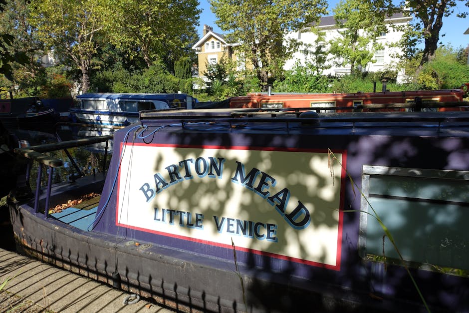 A close-up view of a narrowboat named 'Barton Mead' with a white rectangular signboard on its dark blue hull, featuring bold blue letters indicating its name and location as Little Venice. The boat is