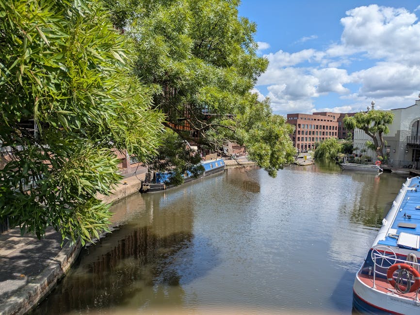 A close-up view of a narrowboat named 'Barton Mead' with a white rectangular signboard on its dark blue hull, featuring bold blue letters indicating its name and location as Little Venice. The boat is