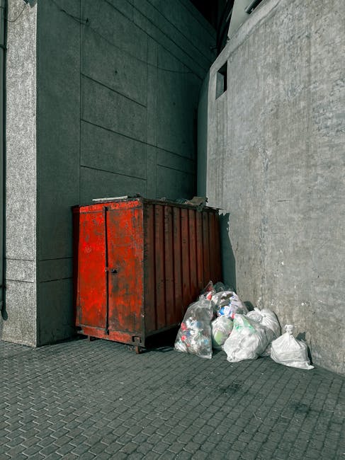 A collection of overflowing rubbish bins and scattered waste bags on a paved sidewalk in front of a commercial building, with various black, red, and grey refuse containers filled with mixed paper, ca